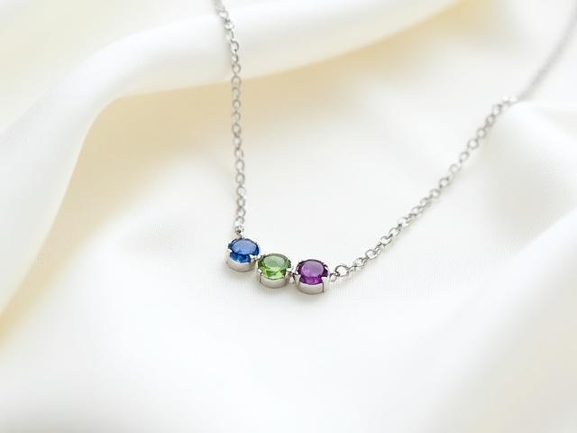 Dainty sterling silver layering necklace with three small, polished birthstones (peridot, amethyst, sapphire) representing a family, on a light fabric backdrop.